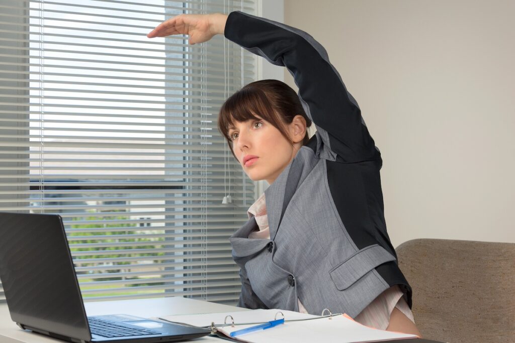 woman doing workouts for busy moms, doing overhead reach stretch while sitting at desk looking at laptop