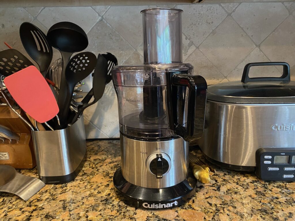 There are so many food processor benefits. Image shows food processor on kitchen counter along with cooking utensils and slow cooker.
