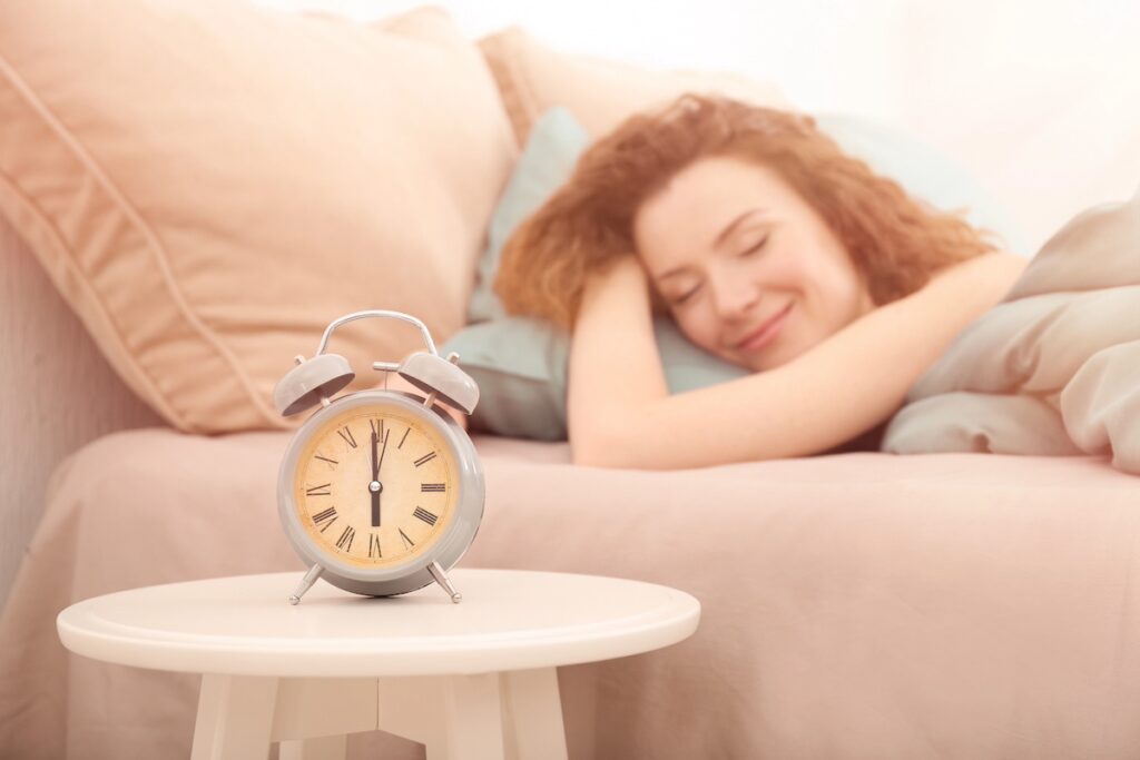 image shows woman who knows how to sleep better, sleeping peacefully in bed with alarm clock next to her