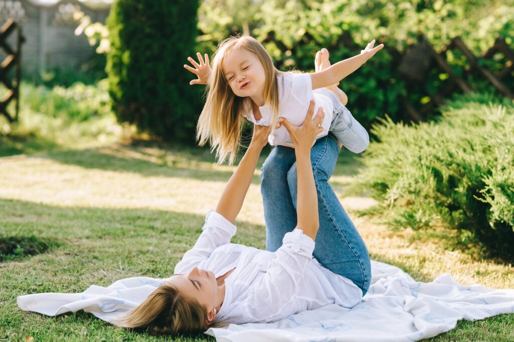 image shows mother laying down on blanket outside with daughter resting on her legs playing airplane, being outdoors is one of the healthy family habits