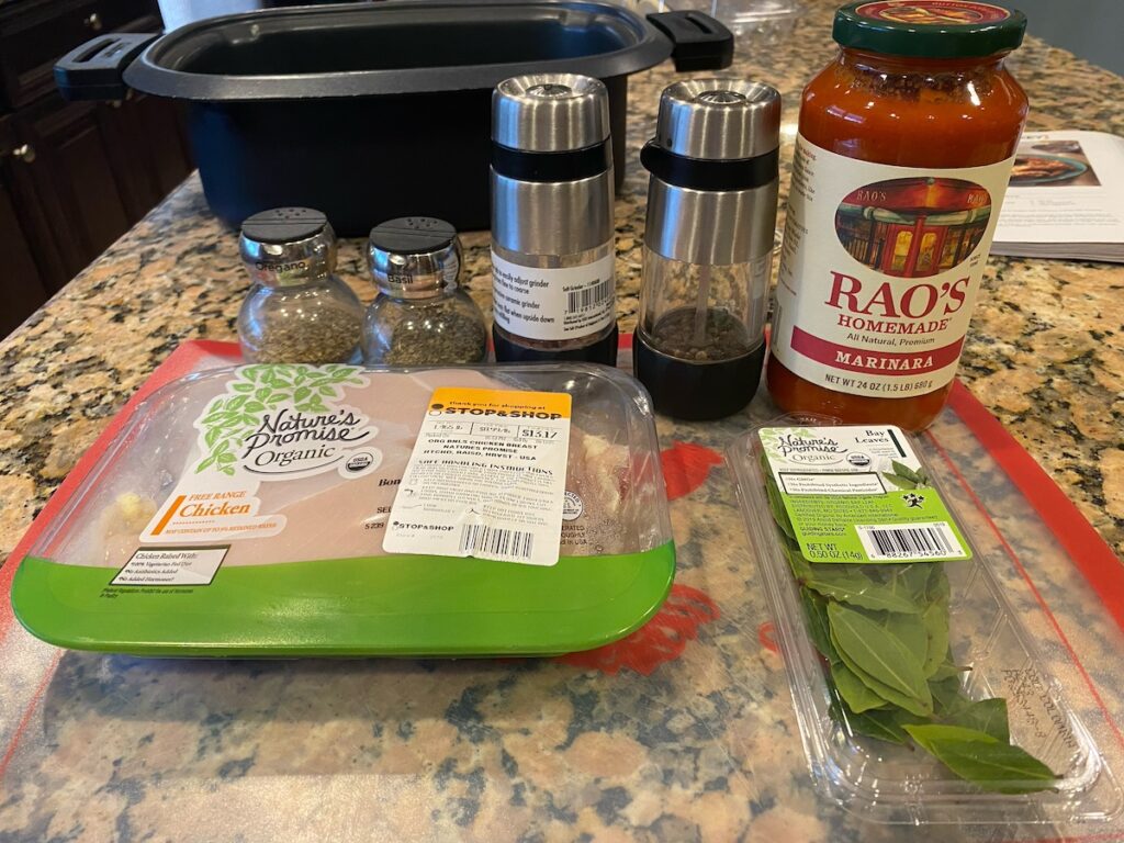 image shows ingredients for slow cooker Italian chicken recipe on kitchen island - chicken breast, bay leaves, basil, oregano, salt, pepper, marinara sauce