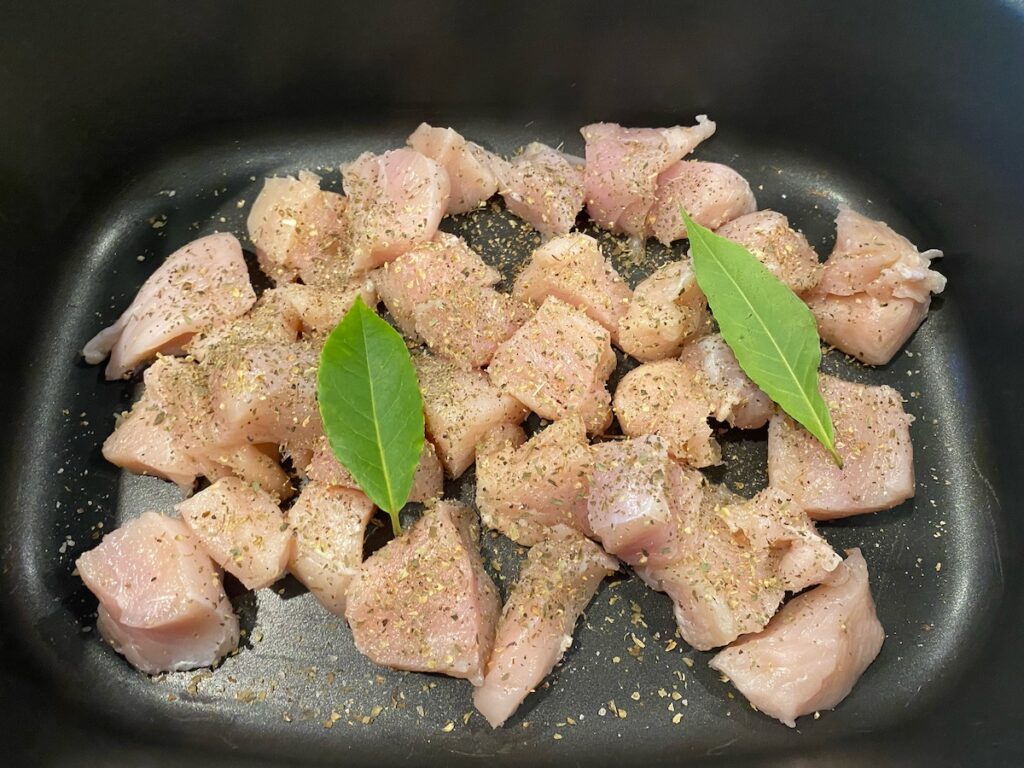 image shows cubed chicken in slow cooker covered in spices and bay leaves