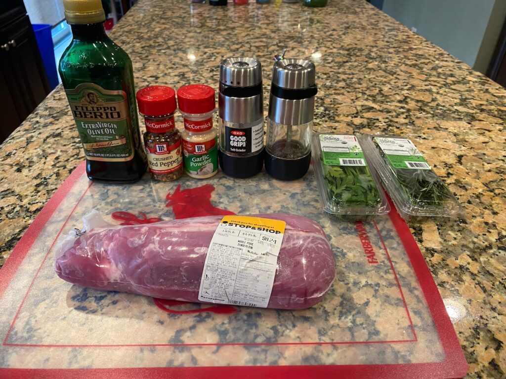 image shows ingredients for the slow cooker herbed pork roast recipe on kitchen island - pork tenderloin, olive oil, ground red pepper, garlic powder, salt, pepper, parsley, and rosemary
