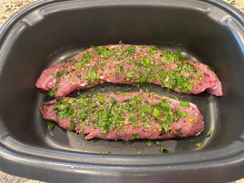 image shows pork tenderloin in slow cooker covered in herbs