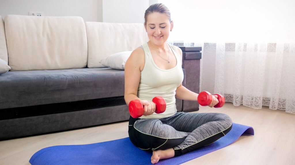 image shows woman smiling on yoga mat in living room, lifting weights, strength training is one of the low-impact workouts for women