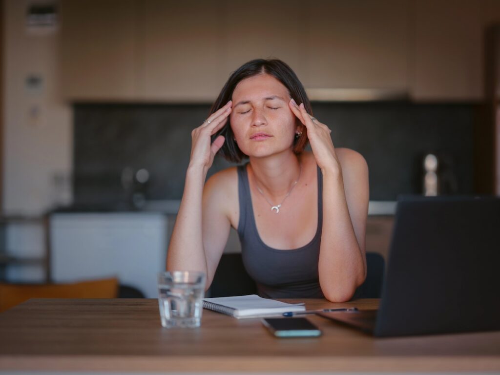 image shows woman in kitchen at home sitting at table, holding her head, suffering from mom burnout