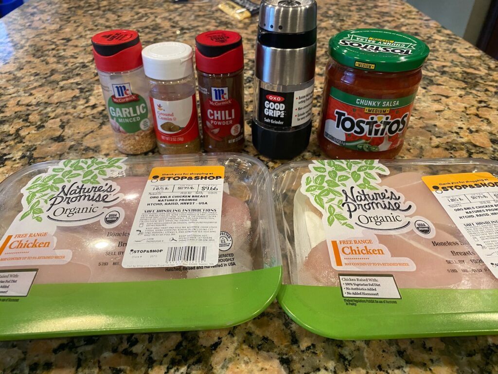 image shows ingredients for healthy slow cooker chicken tacos recipe on kitchen island - chicken, garlic, cumin, chili powder, salt, and salsa