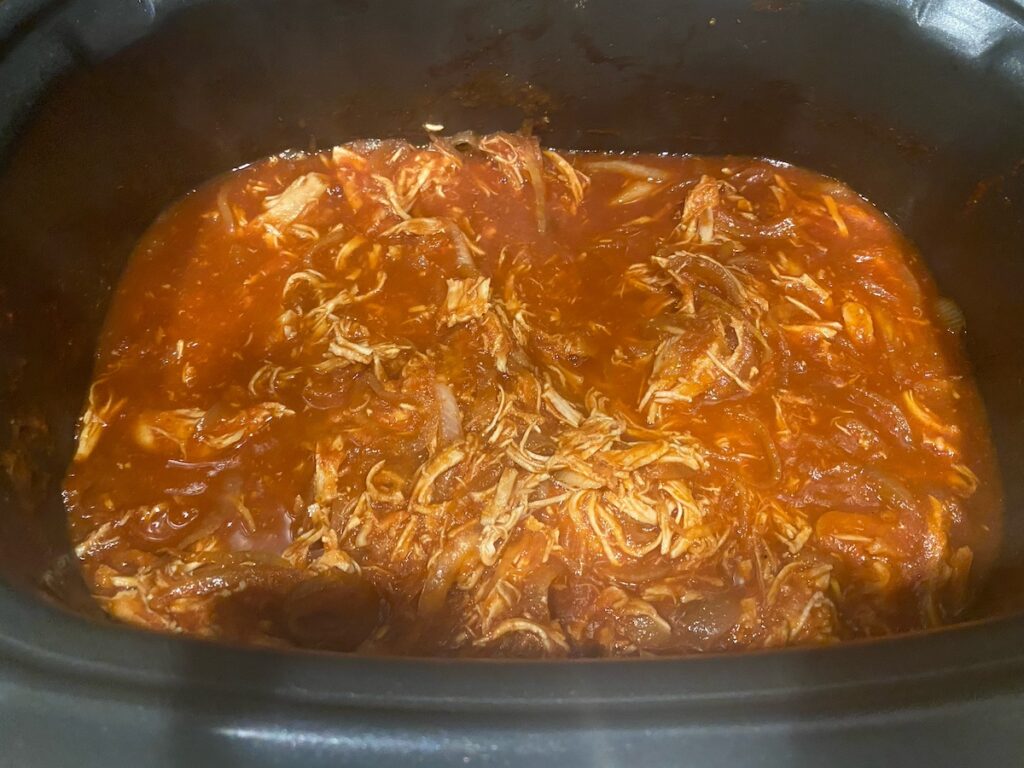 image shows shredded chicken combined with tomato sauce mixture in the slow cooker